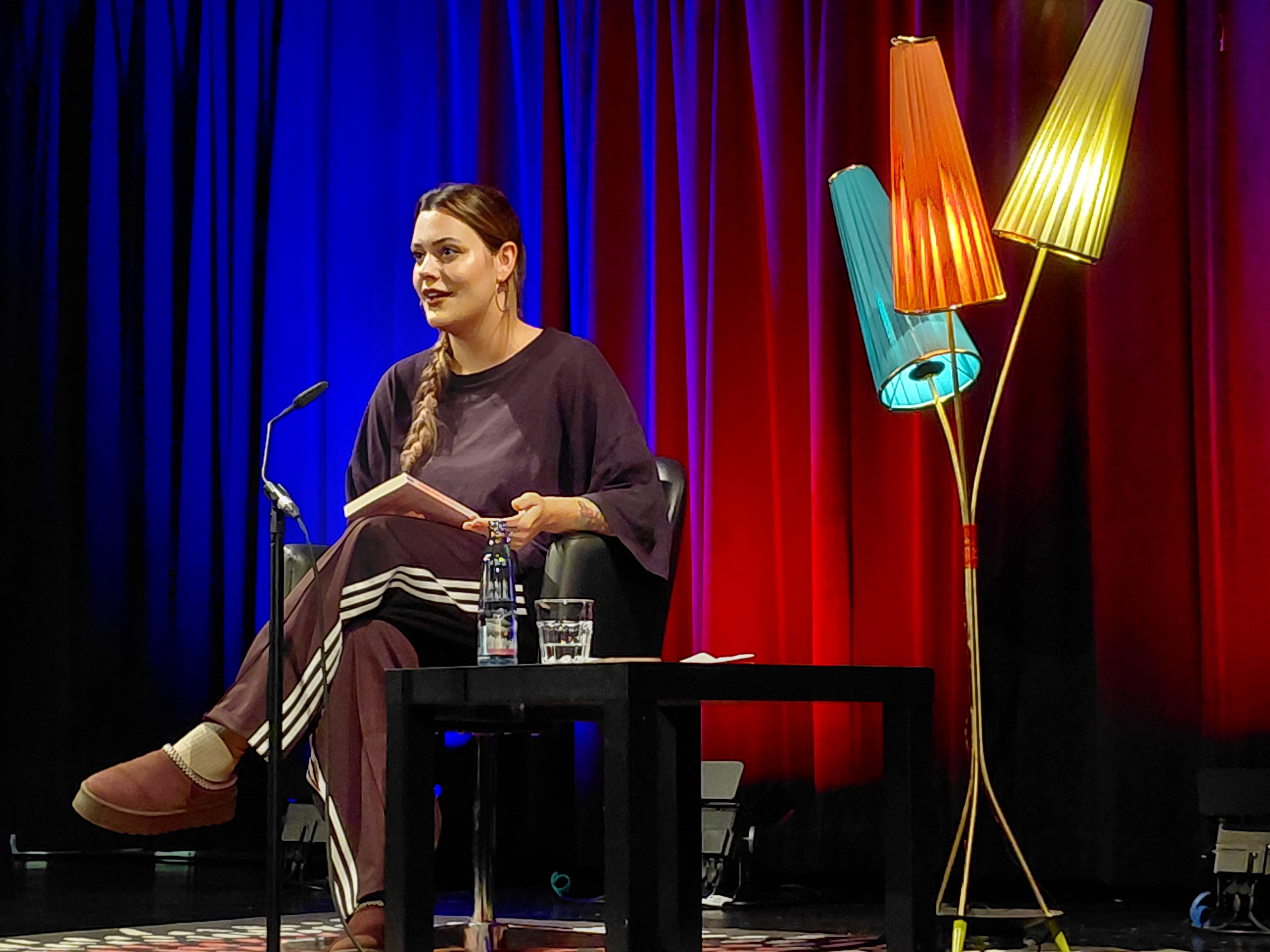 Ein Foto von Joelle Westerfeld auf der Bühne. Links eine Stehlampe mit drei bunten Schirmen. Links Joelle sitzend mit Buch. Hintergrund Vorhang blau und rot beleuchtet.