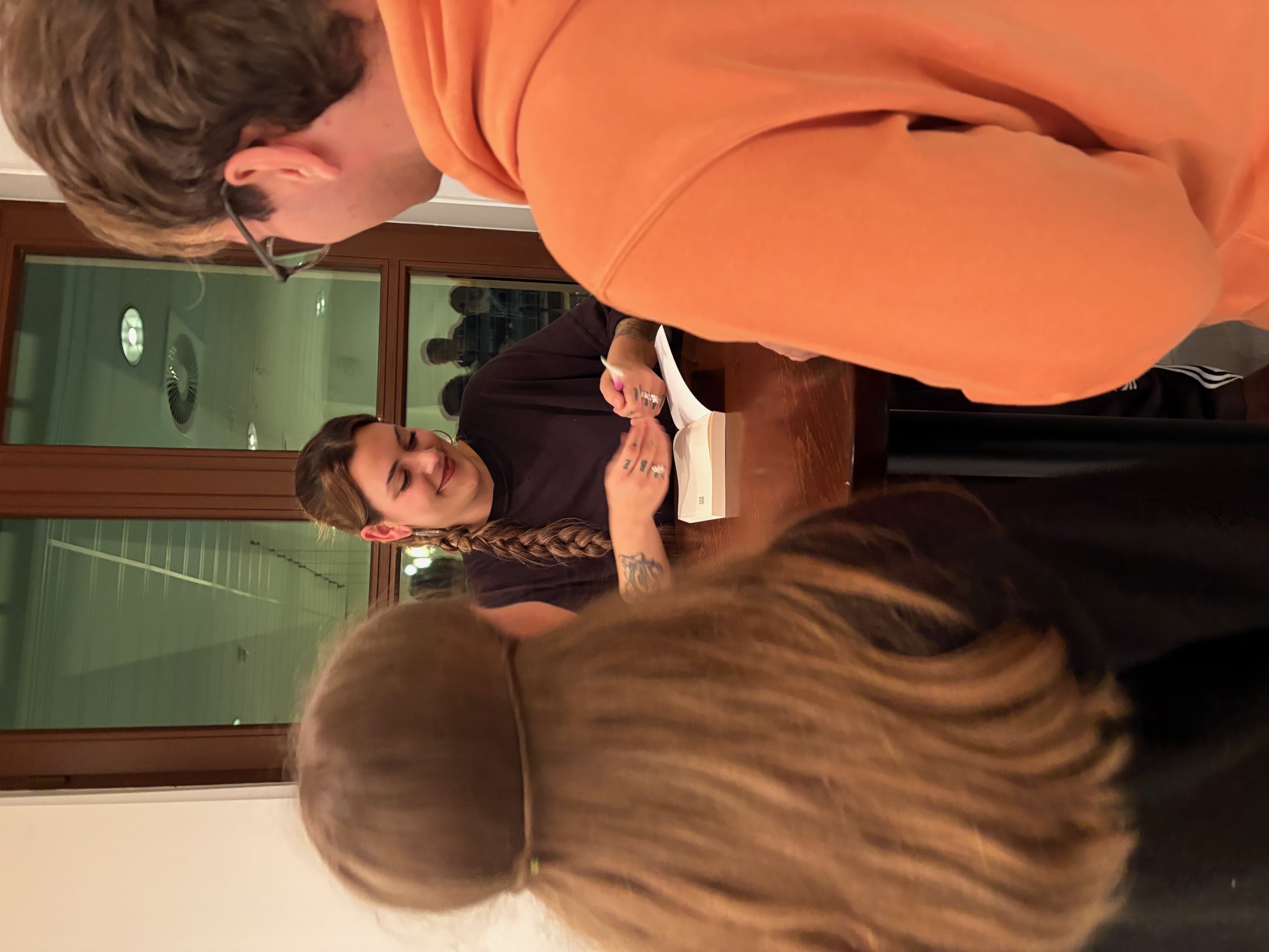 Foto von nach der Lesung. Joelle Westerfeld steht an einem Stehtisch, davor zwei Personen. Sie sind miteinader im Gespräch, Joelle signiert ein Buch.
