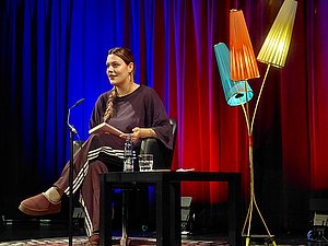 Ein Foto von Joelle Westerfeld auf der Bühne. Links eine Stehlampe mit drei bunten Schirmen. Links Joelle sitzend mit Buch. Hintergrund Vorhang blau und rot beleuchtet.